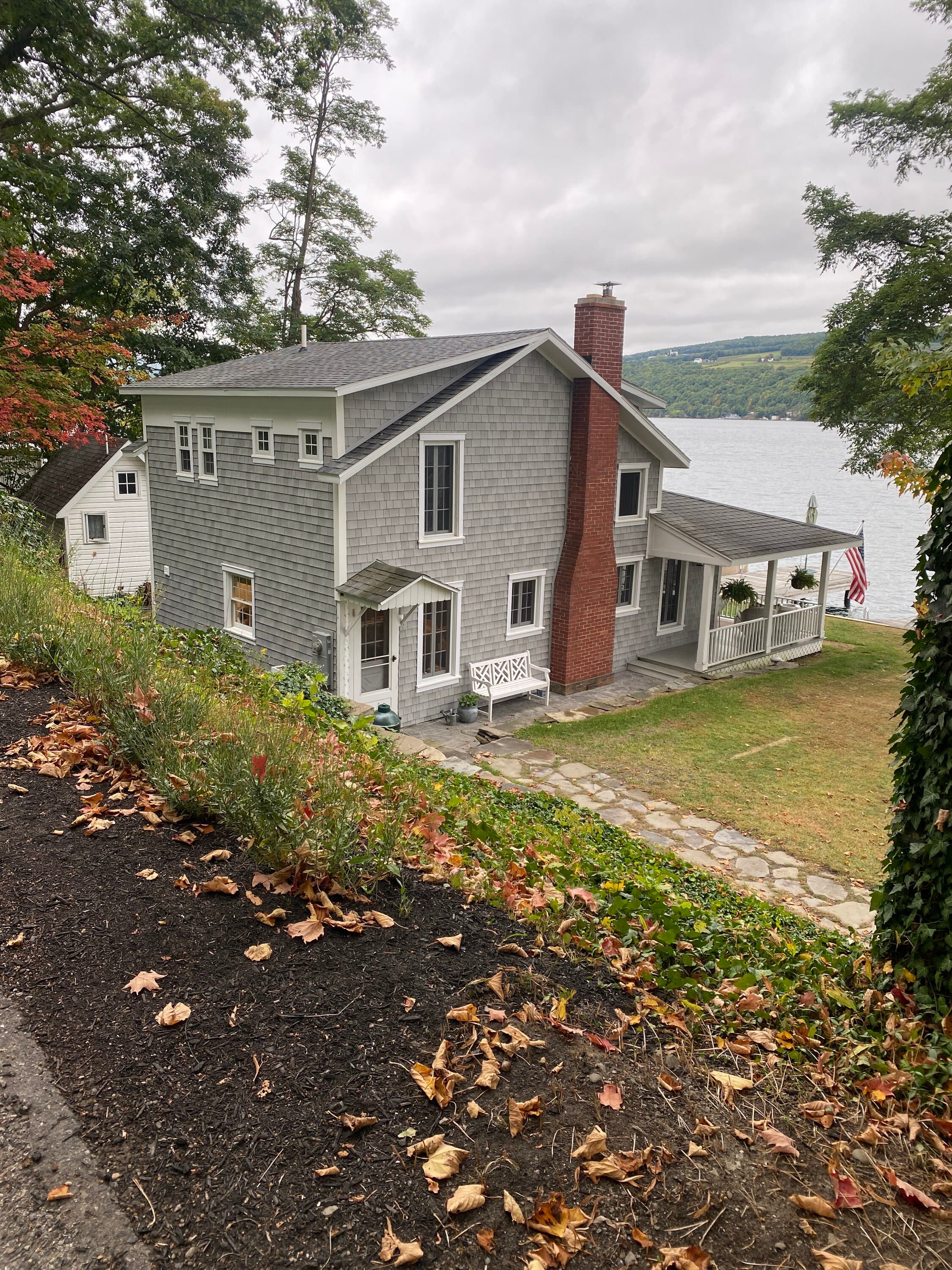 Gray shingled two-story lake house with brick chimney and covered porch overlooking water