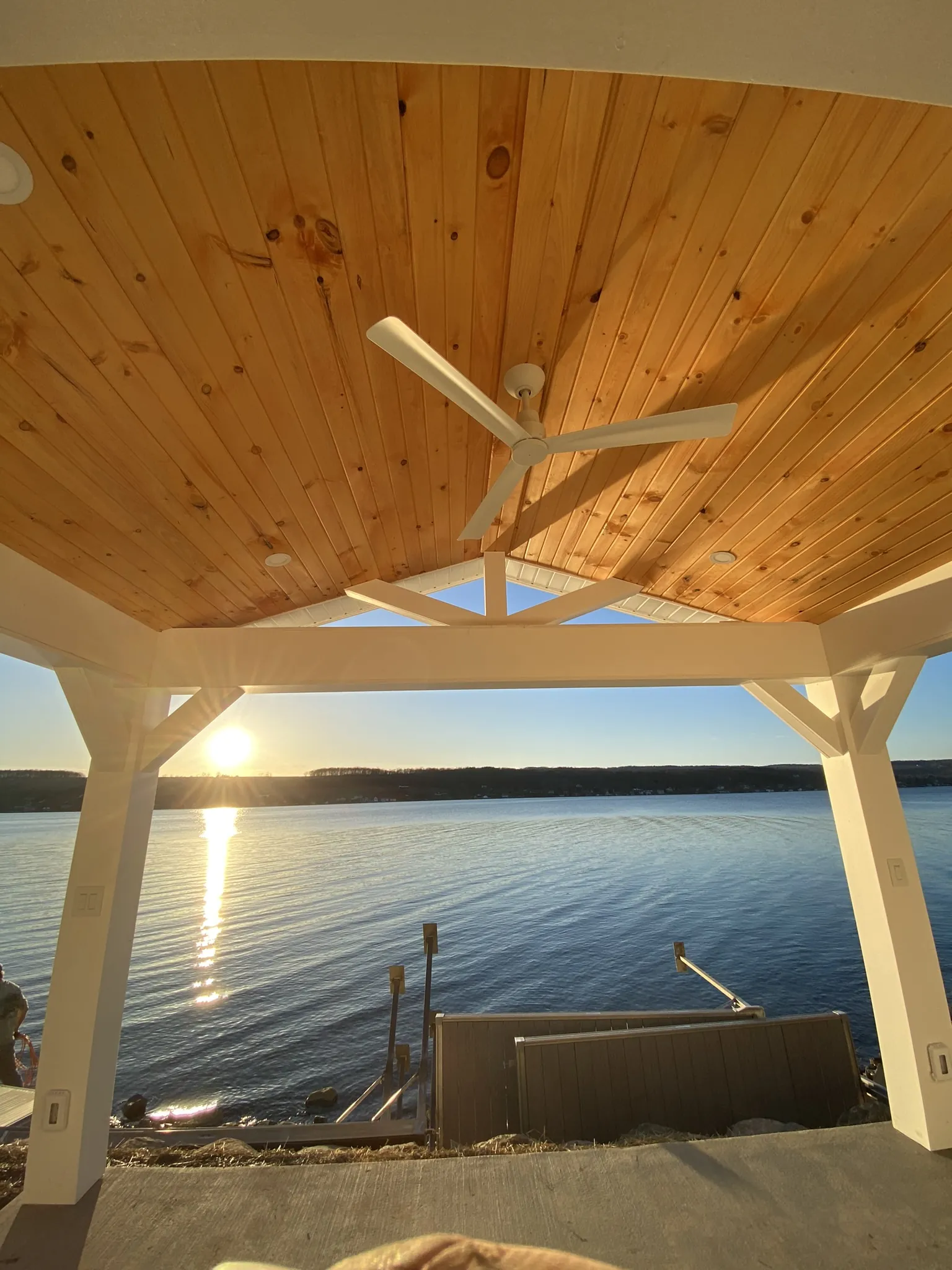 Covered dock with wood ceiling and ceiling fan overlooking calm lake at sunset