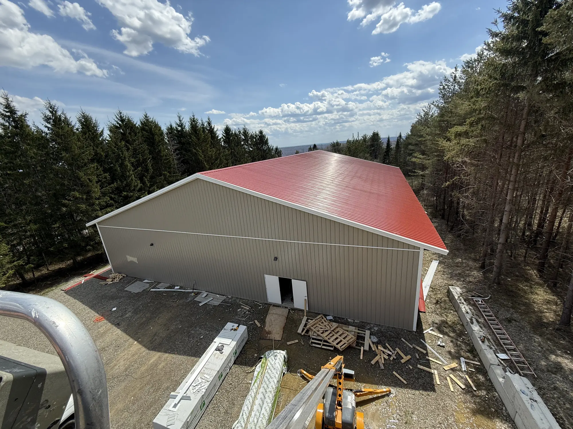 Aerial view of new beige pole barn with red metal roof surrounded by trees